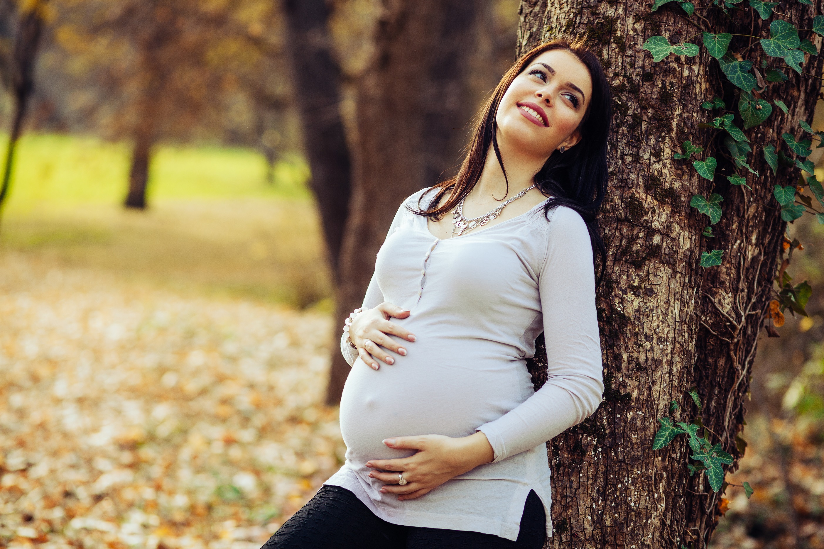 Pregnant woman posing in the park leaning on a tree.